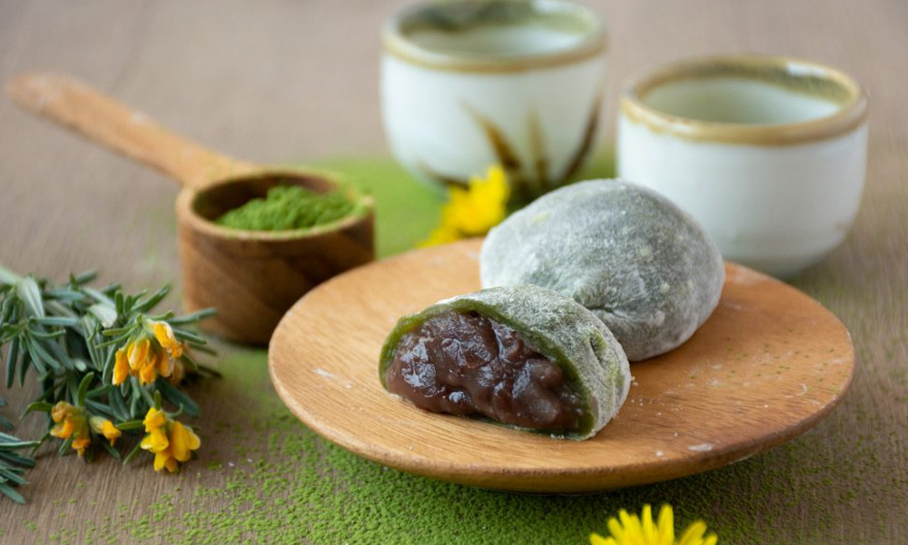 Green mochi balls filled with adzuki red bean paste on a wooden board with matcha tea cups behind the plate